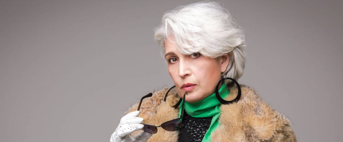 Portrait of senior rich woman holding sunglasses in front of her. Vogue lady in fur coat posing in studio isolated on grey background.