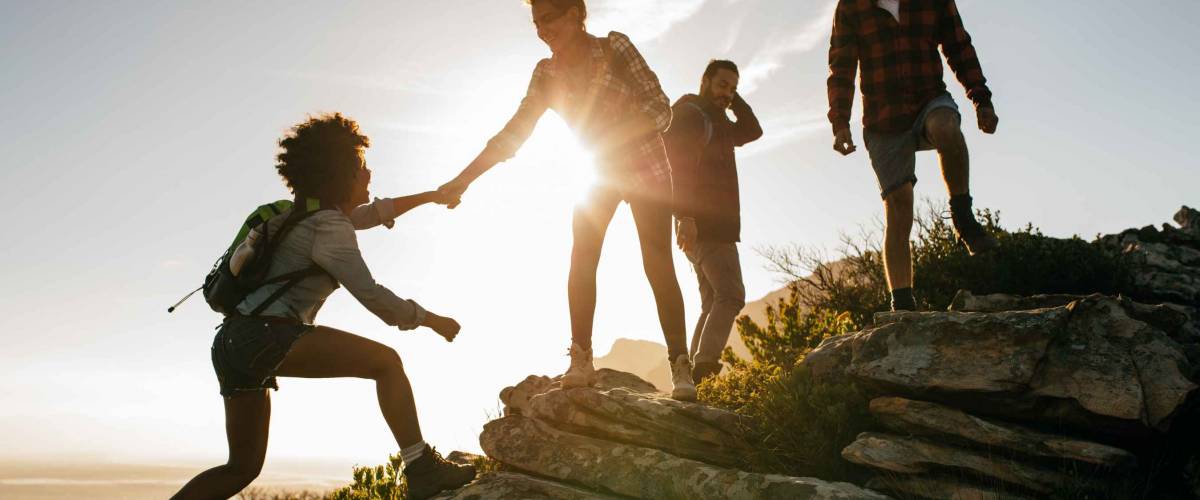 Group of hikers on a mountain. Woman helping her friend to climb a rock. Young people on mountain hike at sunset.