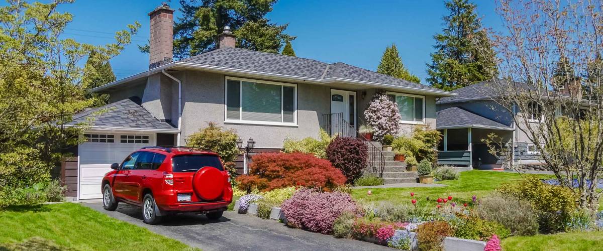 Modest residential house with red car parked on driveway in front. Family house with blossoming flowers on the front yard