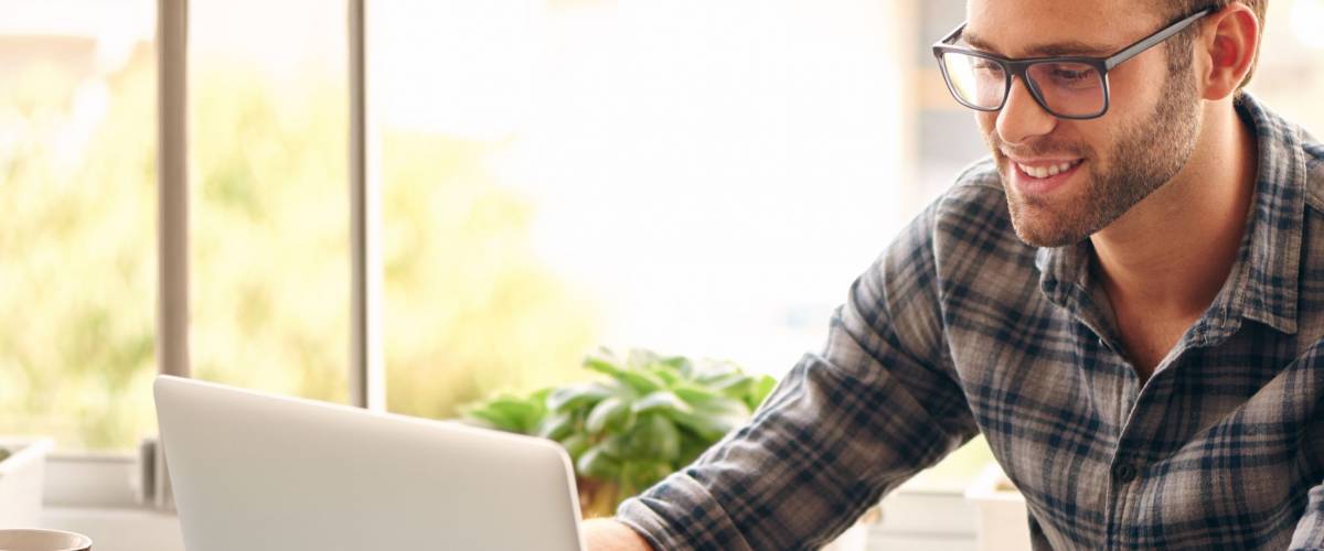 Happy young man, wearing glasses and smiling, as he works on his laptop to get all his business done early in the morning with his cup of coffee
