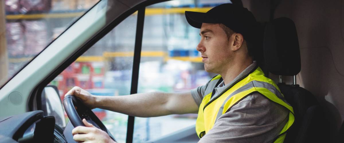 Delivery driver driving van with parcels on seat outside warehouse