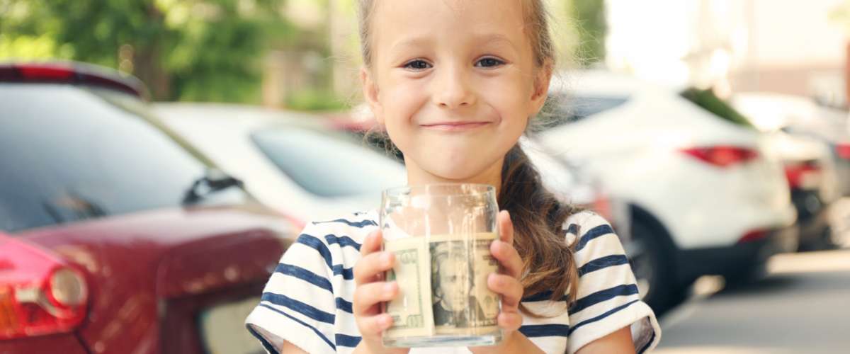 Cute girl holding bank with money in hands and sitting in shopping cart, outdoors