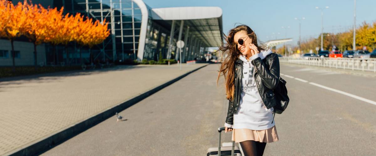 Happy cheerful traveler hipster woman smiling and going with suitcases on the road over airport building in autumn time. Concept of travel, holidays, journey, trip.