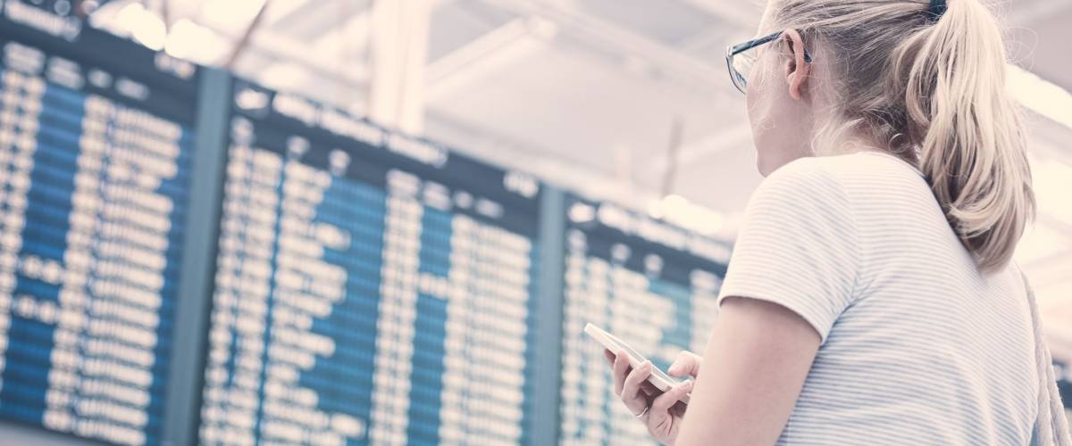 Young blonde woman with phone in her hand and shoulder bag checking flight timetable in international airport - travel concept