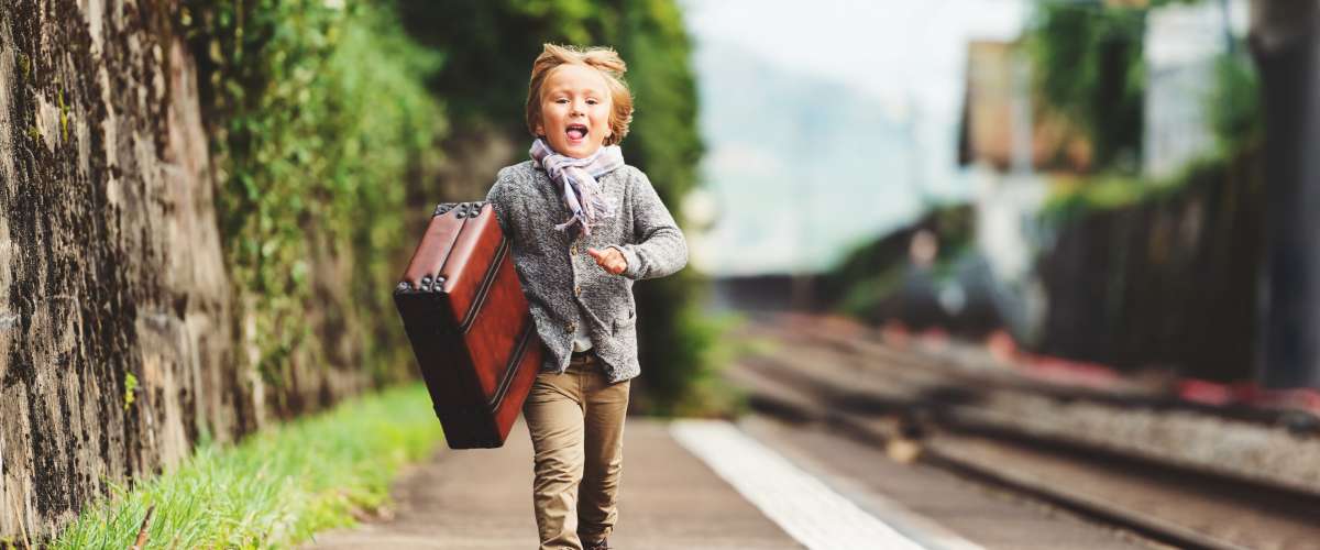 Outdoor portrait of cute little kid boy, run to the train station, wearing beige jacket, trousers, scarf and brown retro shoes, holding old vintage suitcase