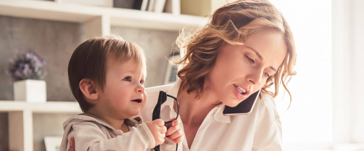 Beautiful business mom is talking on the mobile phone and taking notes while spending time with her cute baby boy at home