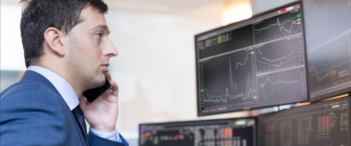 Stock broker trading online while accepting orders by phone. Trader's office with multiple computer screens full of index charts and data analyses.