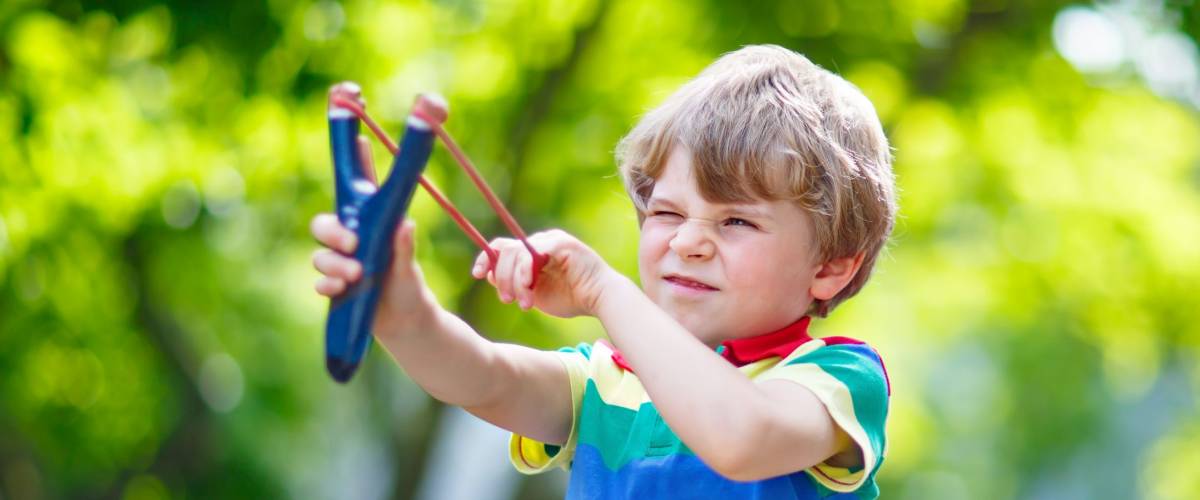 Funny little kid boy shooting wooden slingshot against green tree background. Child having fun in summer. On sunny warm day, active leisure for kids.
