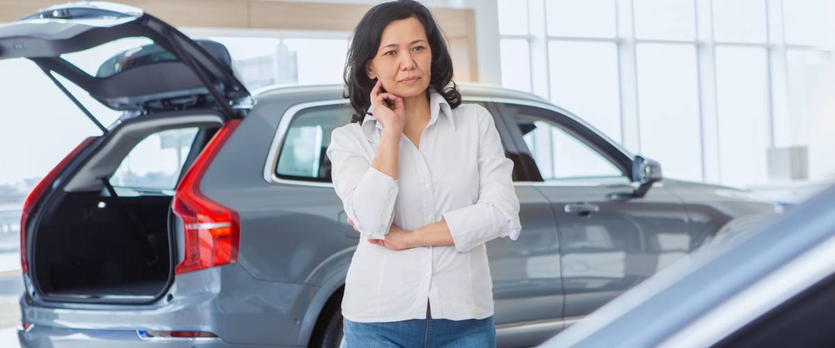 Thoughtful mature Asian woman choosing a car at the dealership. Female customer thinking looking at new cars at the salon copyspace choice deciding buying buyer client consumerism luxury auto