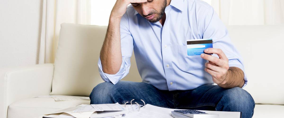 young man at home sitting on sofa with credit card and calculator looking worried or stressed