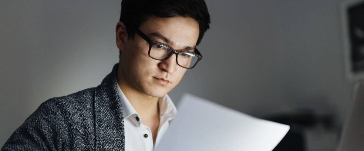 Asian man wearing glasses and casual wear reviewing credit report and working with laptop in dark room late at night, his face lit up by screen