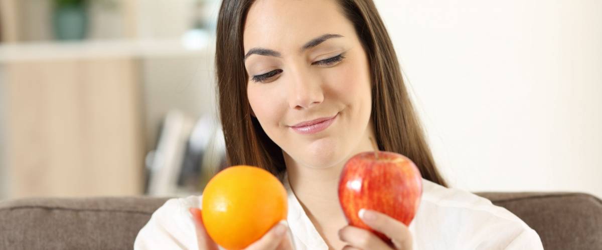 Girl deciding between two fruits an apple and an orange sitting on a couch in the living room at home