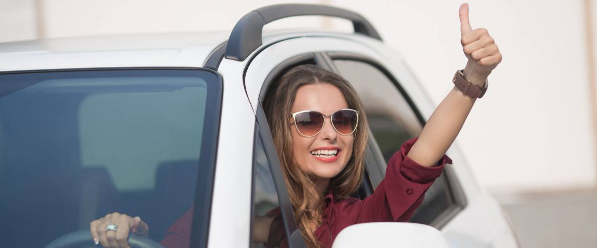 Happy businesslady in dark red shirt driving her car and showing thumb up. Smiling lady in sunglasses taking steering wheel of her new car.