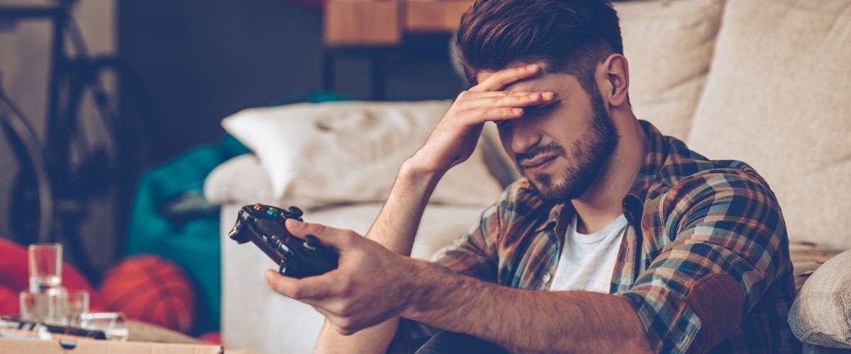 Game over. Frustrated young man holding joystick and keeping his hand on forehead while sitting on the floor in messy room