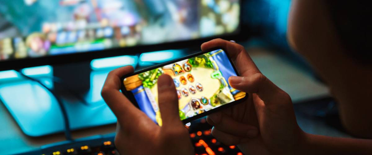 Portrait of teenage gamer boy playing video games on smartphone and computer in dark room wearing headphones and using backlit colorful keyboard