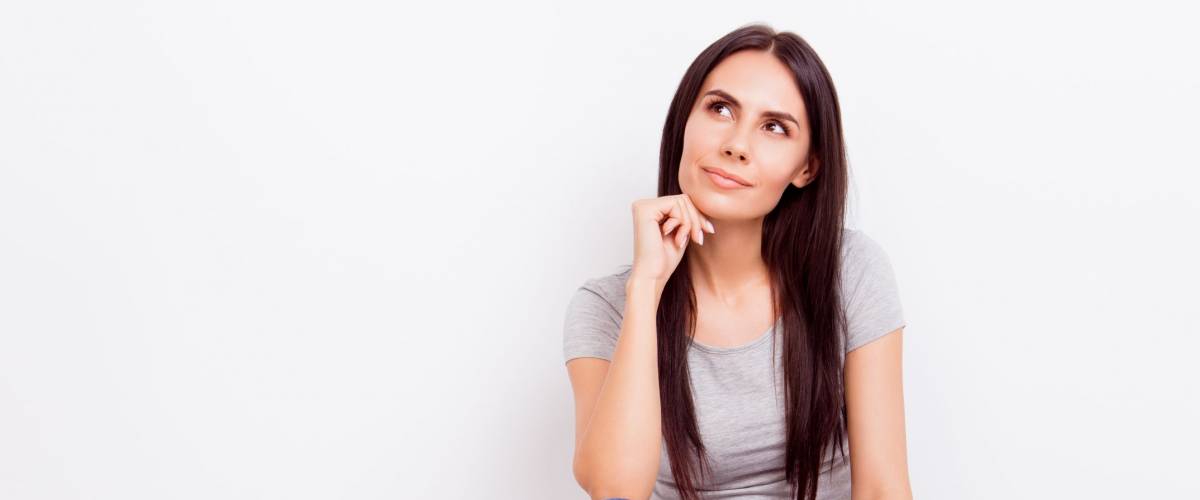Portrait of attractive dreamy pensive brunette lady. She is wearing casual outfit and sitting on the floor with crossed legs on pure white background