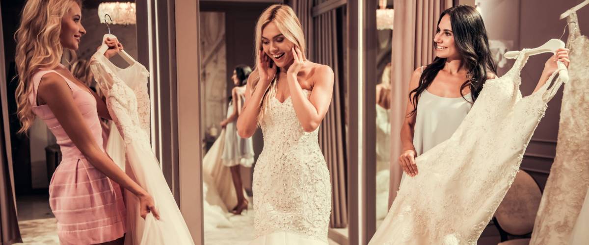 Attractive young bride is smiling while choosing wedding dress in modern wedding salon, girls are showing more dresses