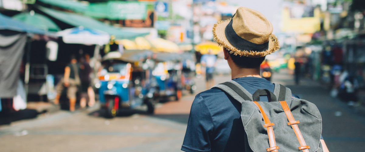 A young traveler walks through a market in Bangkok, Thailand wearing a backpack and a sun hat