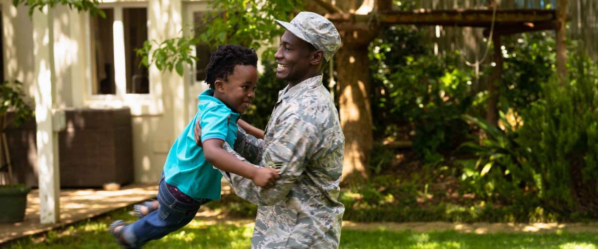 SIde view of a young adult African American male soldier in the garden outside his home, holding his young son and swinging him around, both smiling, their house in the background