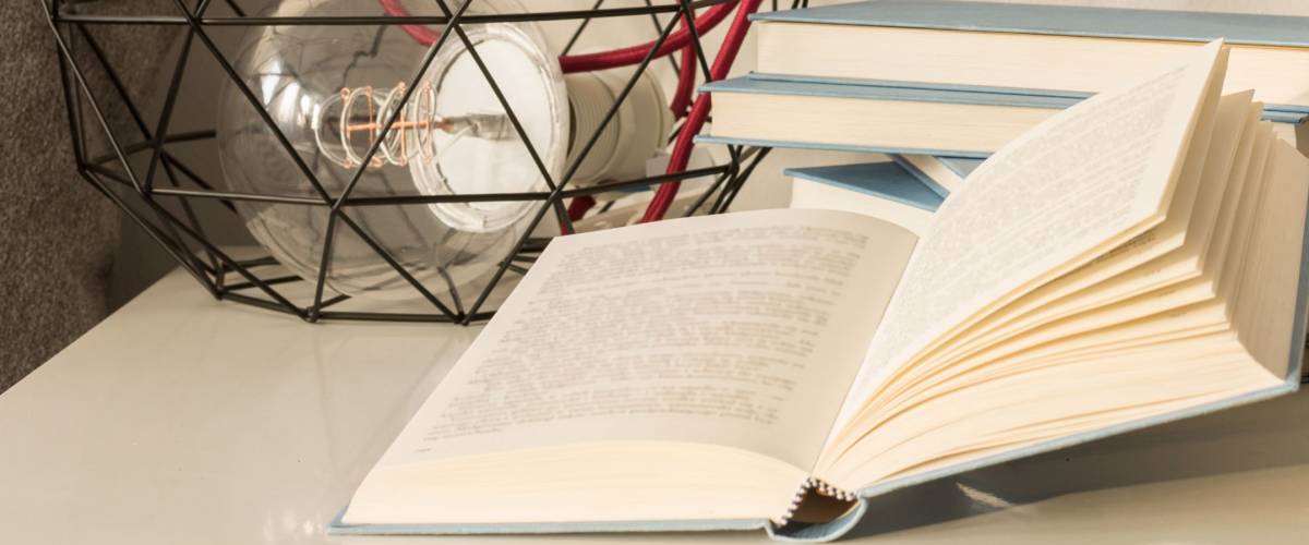 White nightstand with a pile of books in blue cover, one of which is lying open.