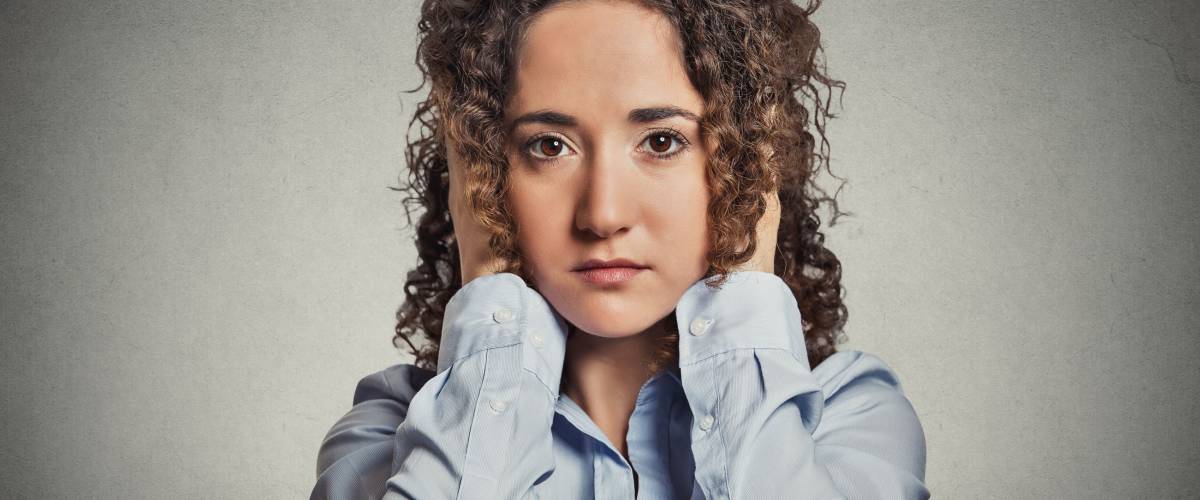 Young woman covering her ears eyes opened isolated grey office wall background. Hear no evil concept idea.