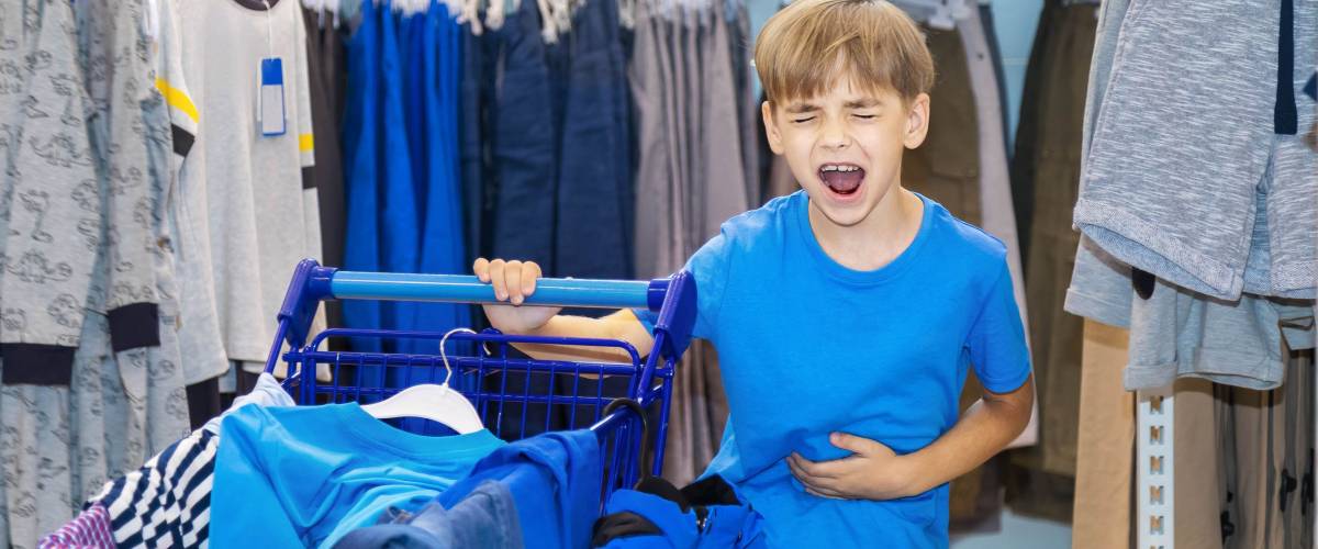 Caucasian boy chooses in shop clothes by new academic year. Is tired, angry and shouts from indignation. Before him the cart loaded with the chosen clothes and footwear