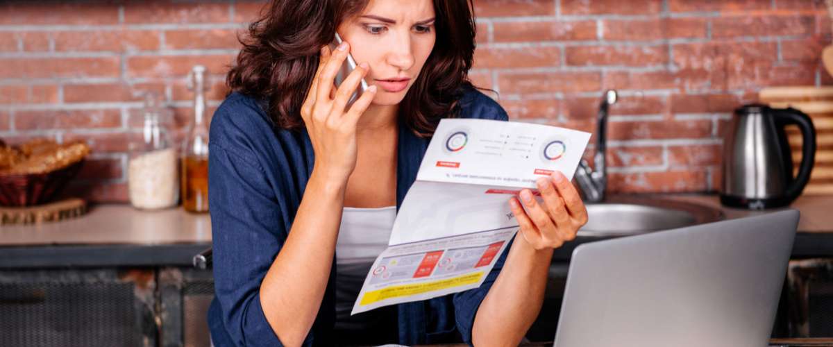 Young woman sitting in the kitchen with dissatisfied face in front of laptop holding utility bills, talking on mobile phone